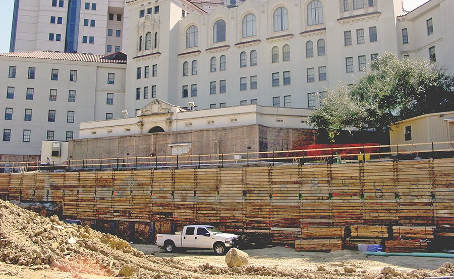 A white pickup truck is parked in front of a construction site with stacked wooden planks and dirt piles, set against a large, multi-story historic building in the background.