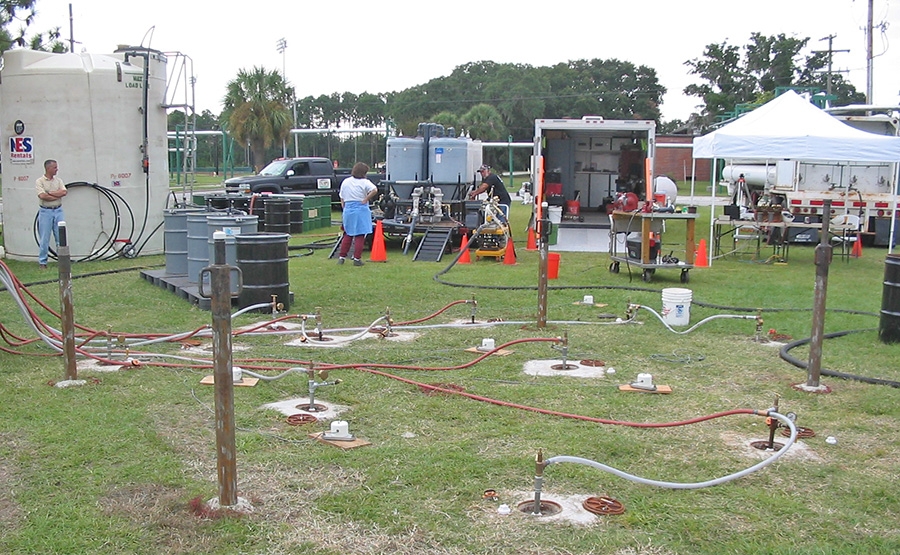 346cfc4cda7a853e9a165b7df6f7b442 Several pipes and hoses connected to wells are set up on a grassy field, with large tanks, barrels, a truck, and workers under tents and by equipment in the background, suggesting an environmental cleanup operation.
