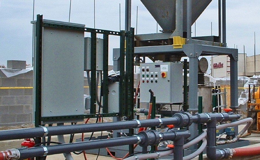 Industrial equipment with an open control panel and several pipes and valves connected below, located outdoors at a construction or manufacturing site. Control buttons and indicators are visible on the panel.