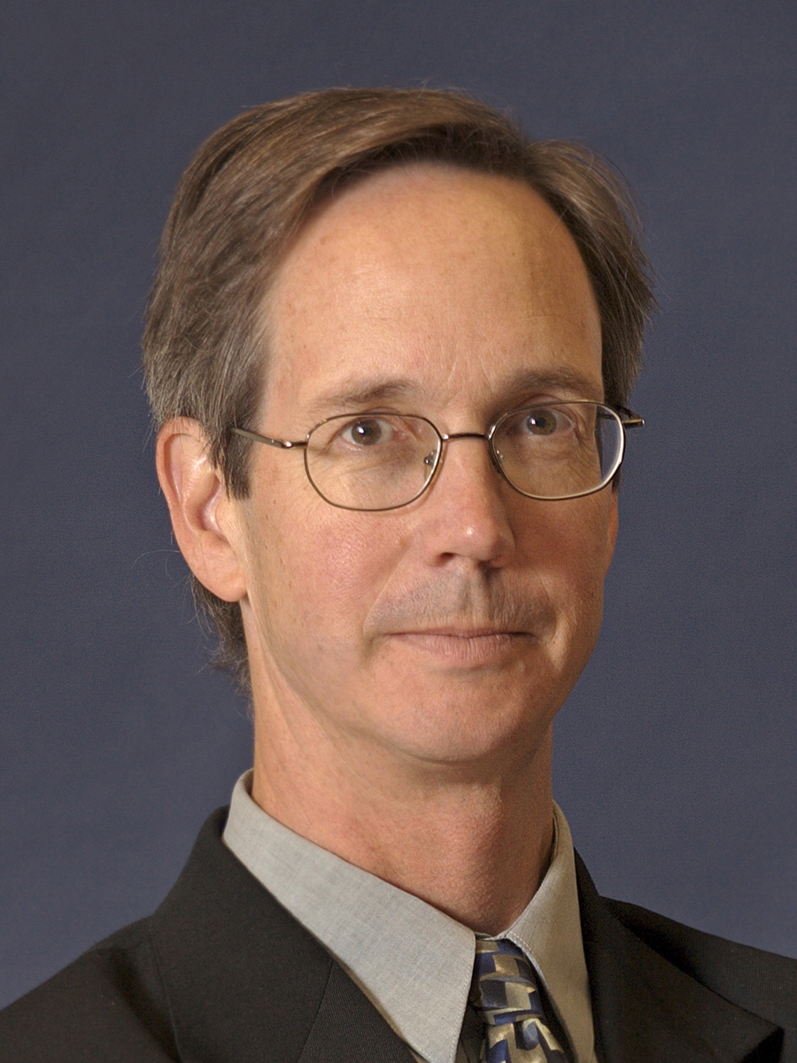 A middle-aged man with short brown hair and glasses wearing a suit, light gray shirt, and patterned tie, posing in front of a plain dark blue background.