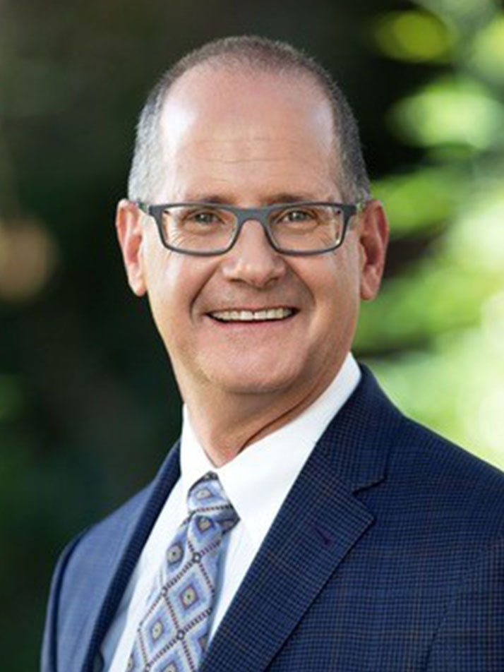 A middle-aged man with short hair and glasses, wearing a dark blue suit, white shirt, and patterned tie, smiles while standing outdoors with greenery in the blurred background.