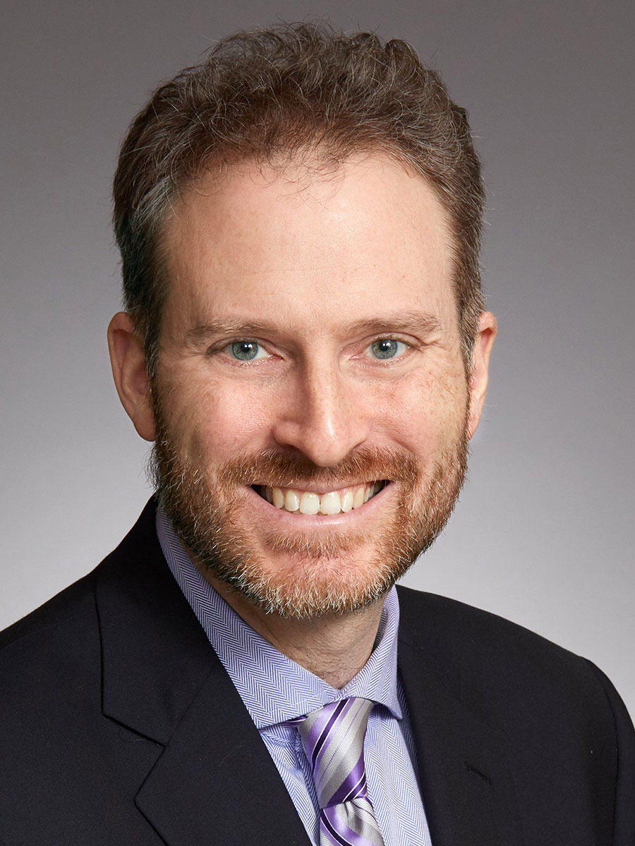 A man with short, light brown hair and a beard is smiling at the camera. He is wearing a dark suit jacket, a light blue shirt, and a striped tie, posed against a plain gray background.