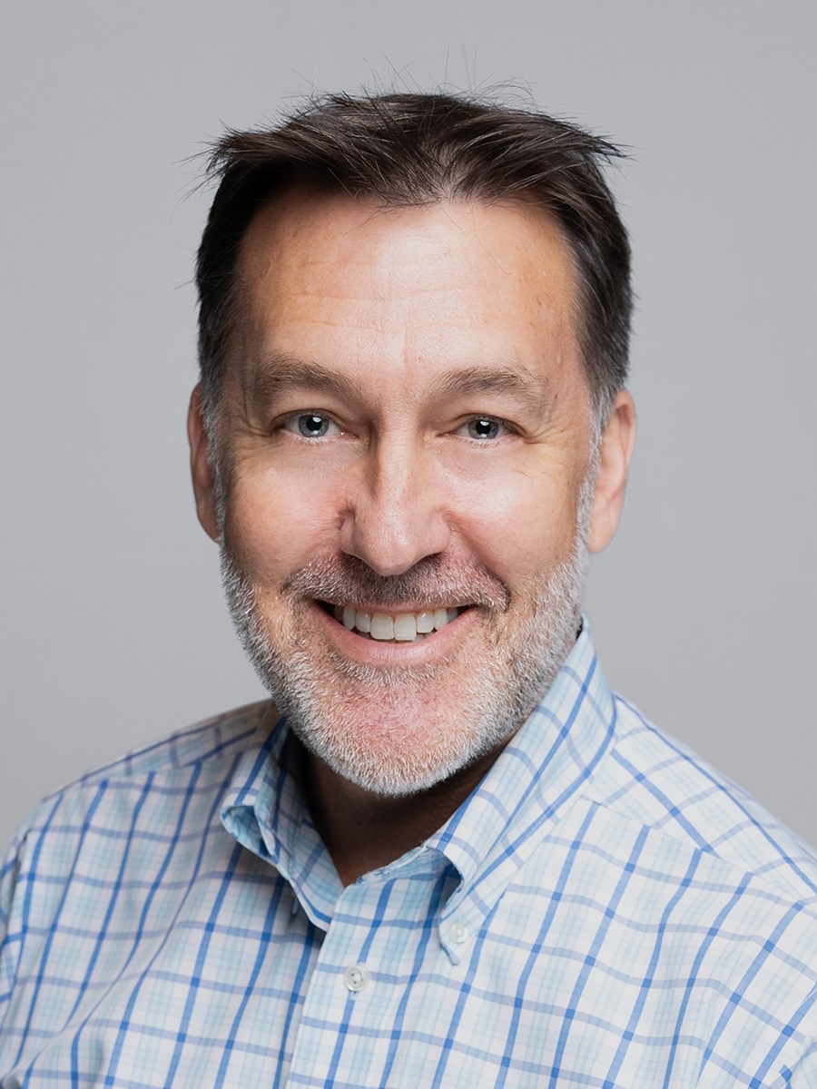 Smiling middle-aged man, identified as Scott Graves, with short brown hair and a salt-and-pepper beard, wears a blue and white checkered shirt while posing against a plain gray background.