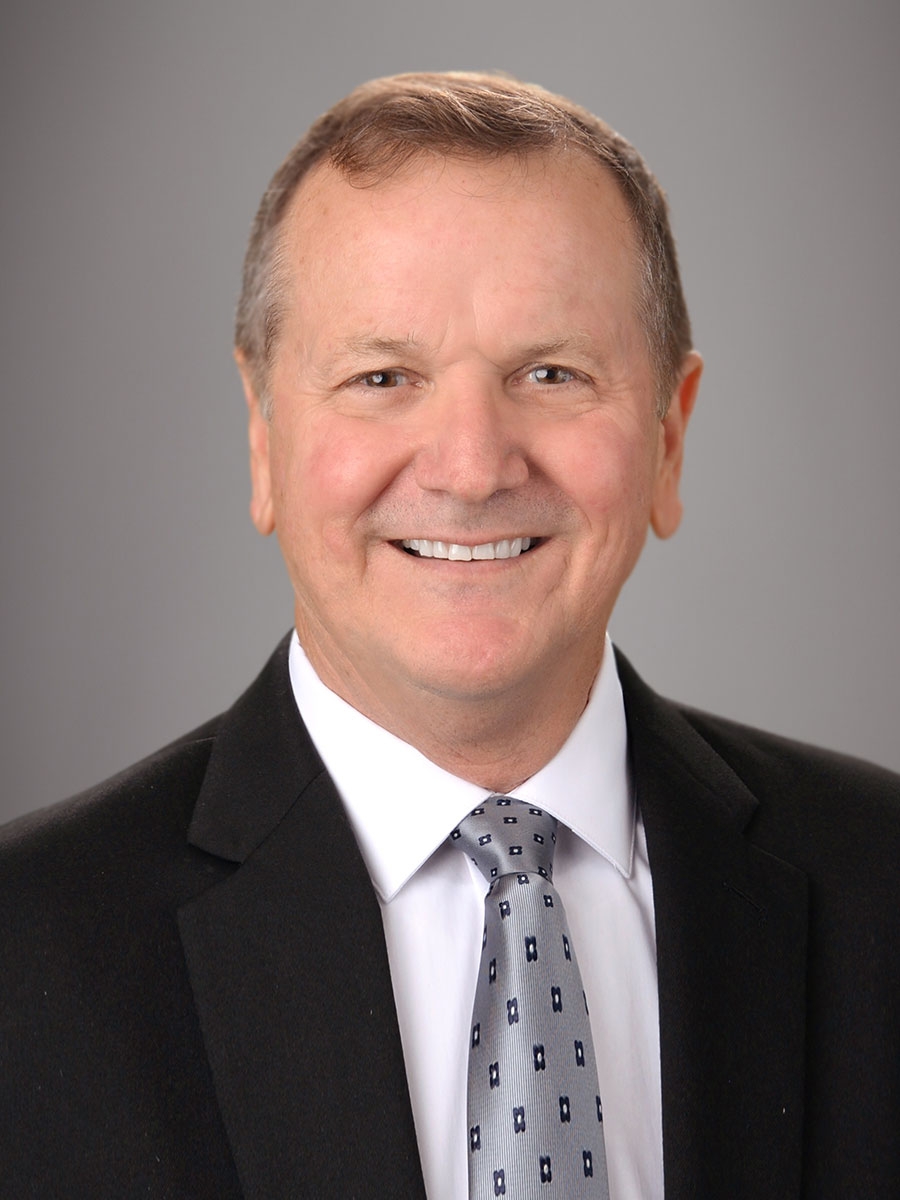 A middle-aged man with short brown hair is smiling and wearing a black suit jacket, white shirt, and patterned gray tie against a plain gray background.