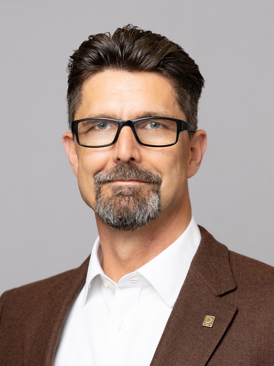 A man with short, dark hair and a gray goatee, wears black glasses, a white shirt, and a brown blazer with a lapel pin as he poses against a plain, light gray background.