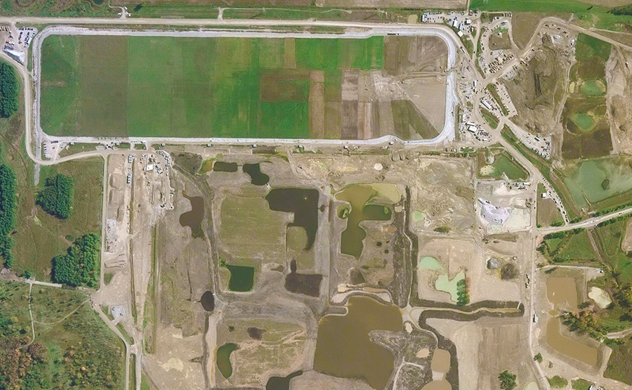 Aerial view of a construction site with rectangular green fields at the top, earthworks, muddy areas, scattered ponds, and surrounding roads and vehicles. The landscape shows ongoing land development.