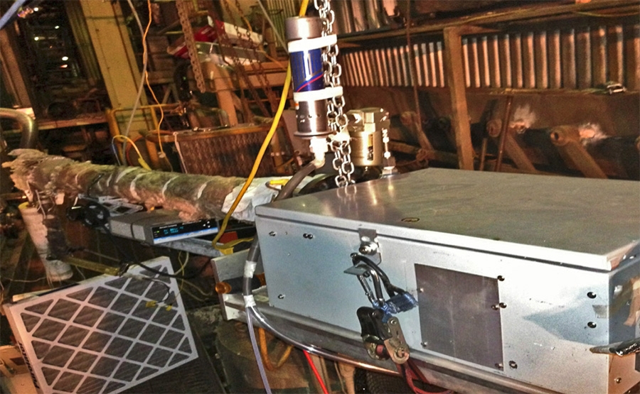 A metal industrial machine with wires and pipes connected, located in a workshop or factory. A can of Red Bull energy drink is suspended above the machine with a chain. The setting appears cluttered and mechanical.