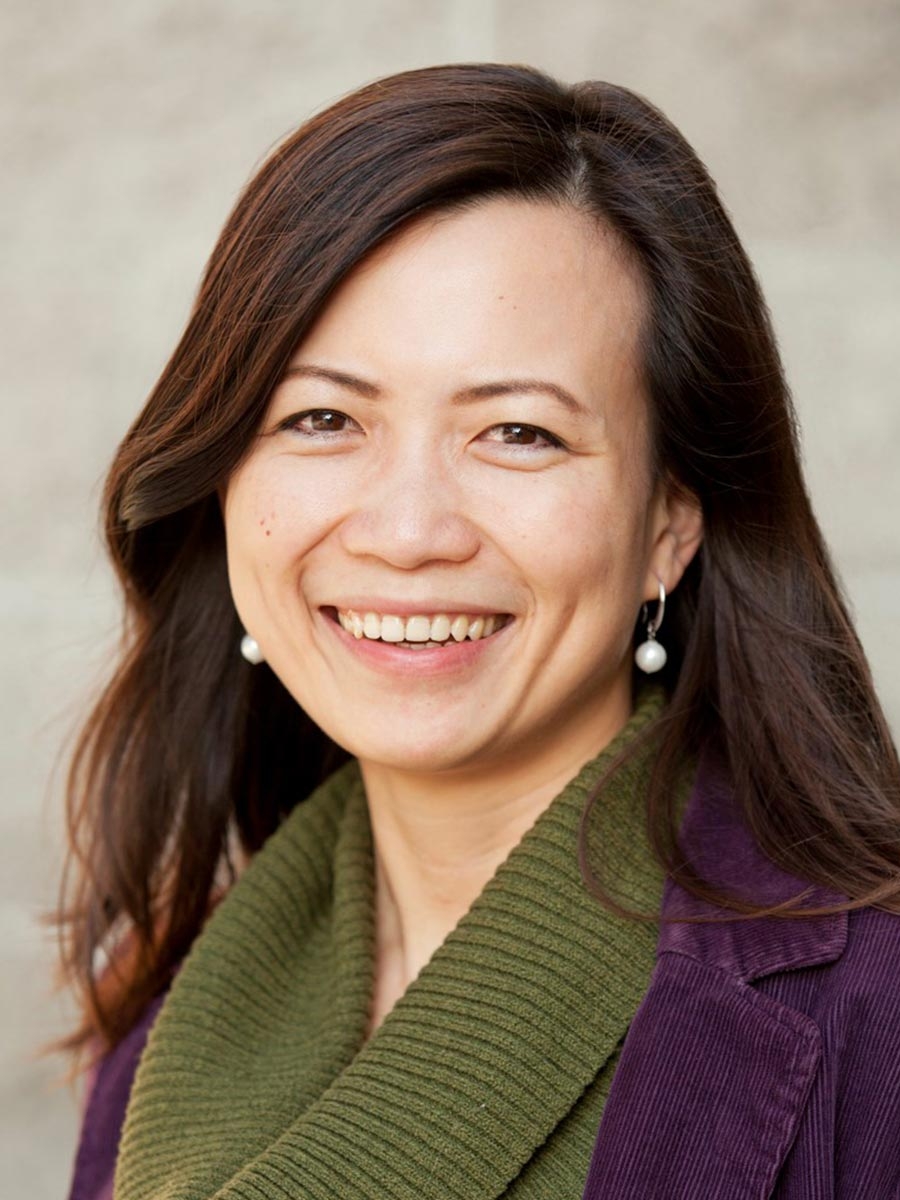 3f4ff99be4325c3c6225fadb049491f0 A woman with long brown hair smiles at the camera. She is wearing pearl earrings, a green scarf, and a purple jacket, with a blurred neutral background.