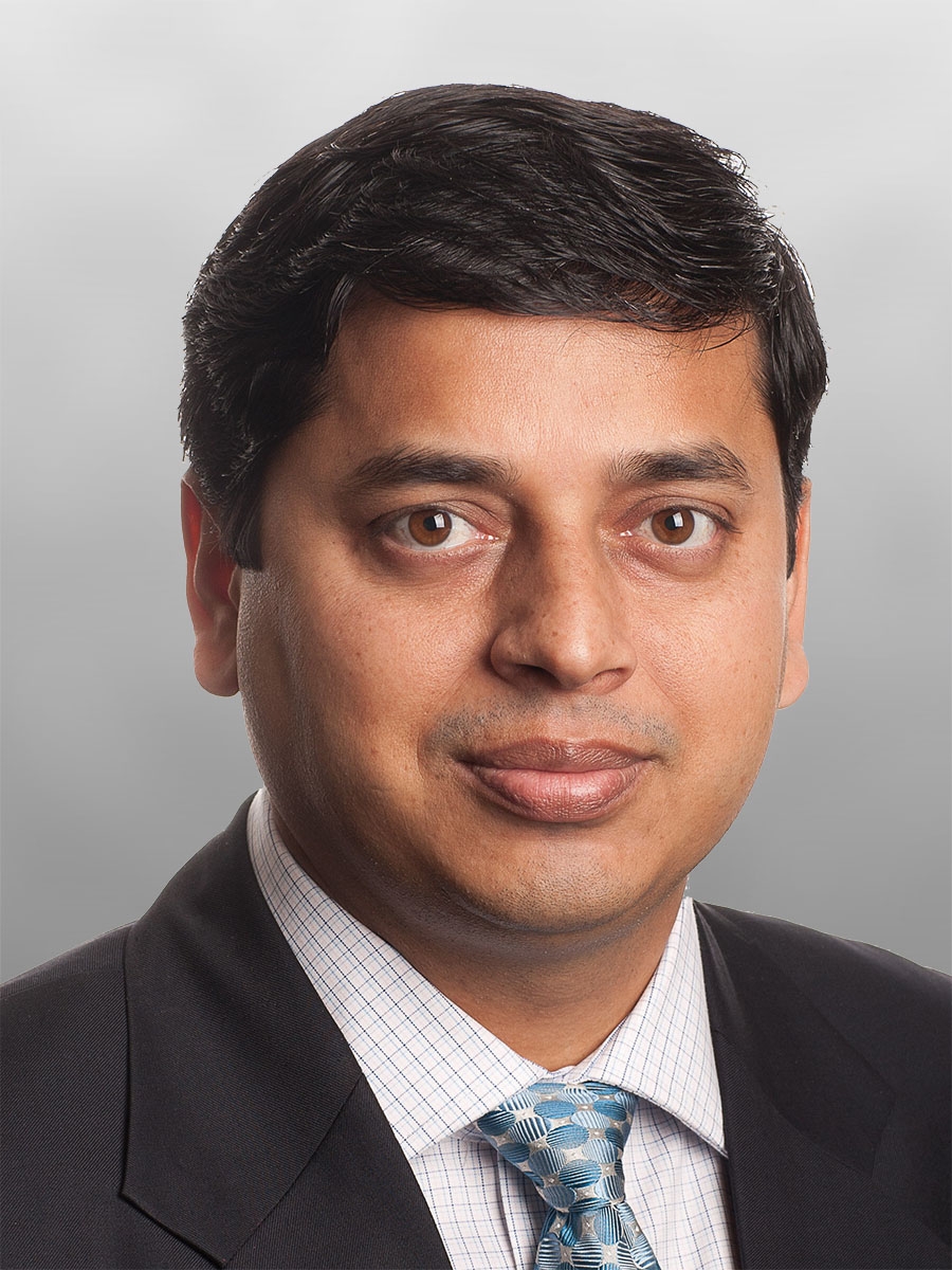 A man with short dark hair wearing a dark suit, light blue patterned tie, and a checkered dress shirt, poses for a professional headshot against a plain light gray background.