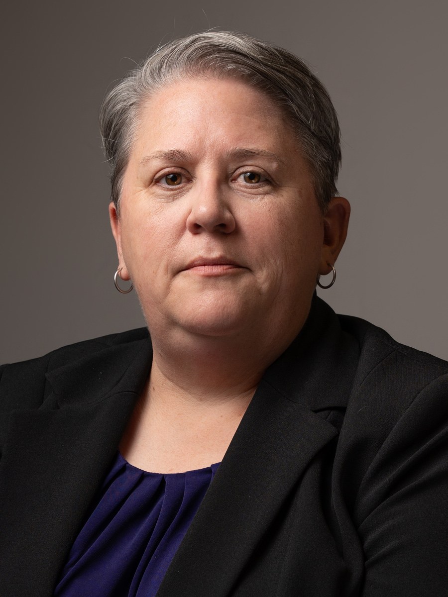 A woman wearing hoop earrings, a black blazer, and a purple top, poses against a plain, neutral background with a calm, serious expression.