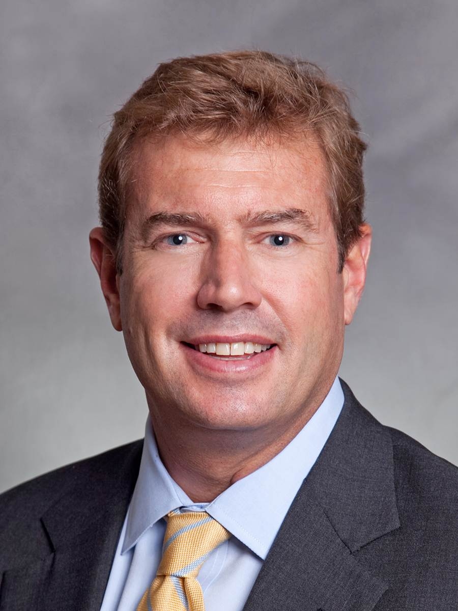 A man with short light brown hair is wearing a gray suit, light blue shirt, and yellow tie, smiling in a professional headshot against a gray background.
