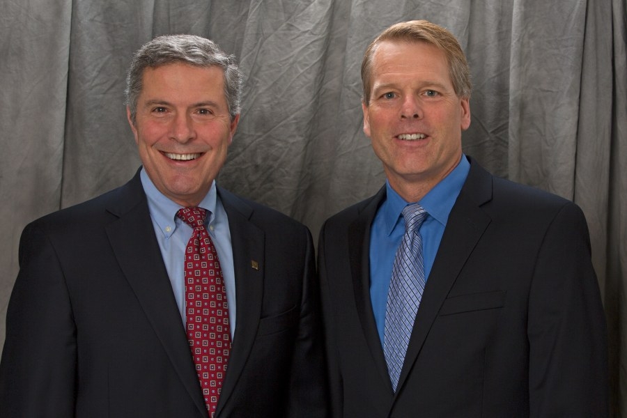 f9ed36389057c757d825df002515e553 Two men in suits are standing side by side, smiling at the camera. The background is a gray, textured curtain. Both men have short hair; one wears a red patterned tie, the other a light blue tie.