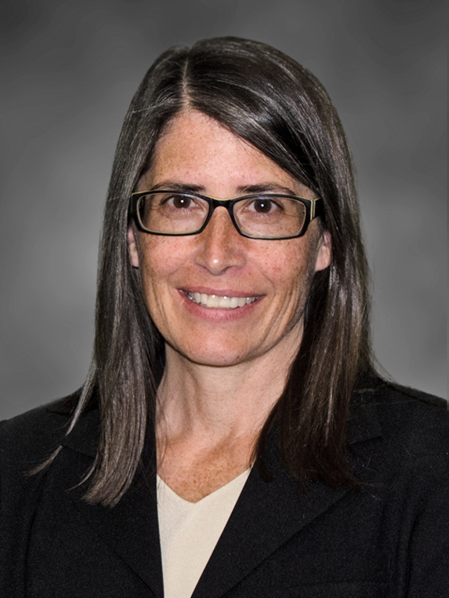 A woman with straight brown hair, wearing glasses, a black blazer, and a light-colored top, smiles in front of a gray background.