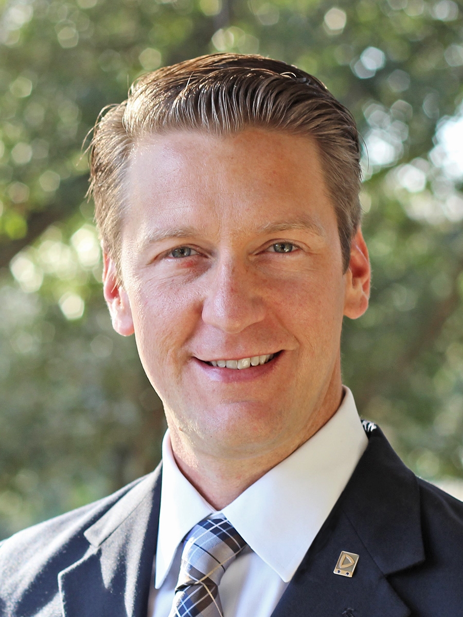 A man in a suit and tie smiles at the camera outdoors, with sunlight filtering through green trees in the blurred background.