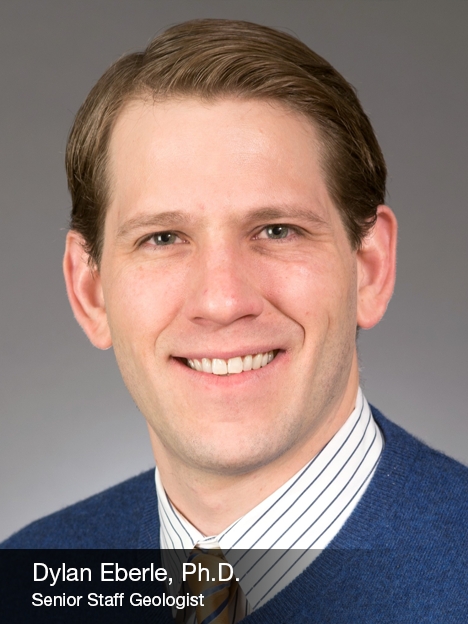 A man with short, light brown hair smiles at the camera, wearing a blue sweater over a striped shirt and tie. Text overlay reads: Dylan Eberle, Ph.D. Senior Staff Geologist.