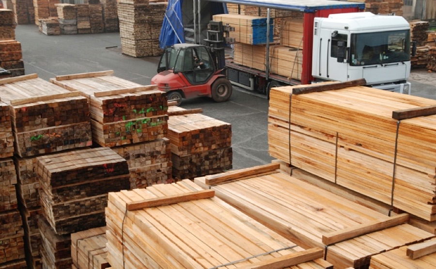 Stacks of cut lumber are piled in a lumberyard. A forklift loads wooden planks onto a flatbed truck, while more stacks of wood are visible in the background. The scene is outdoors and industrial.