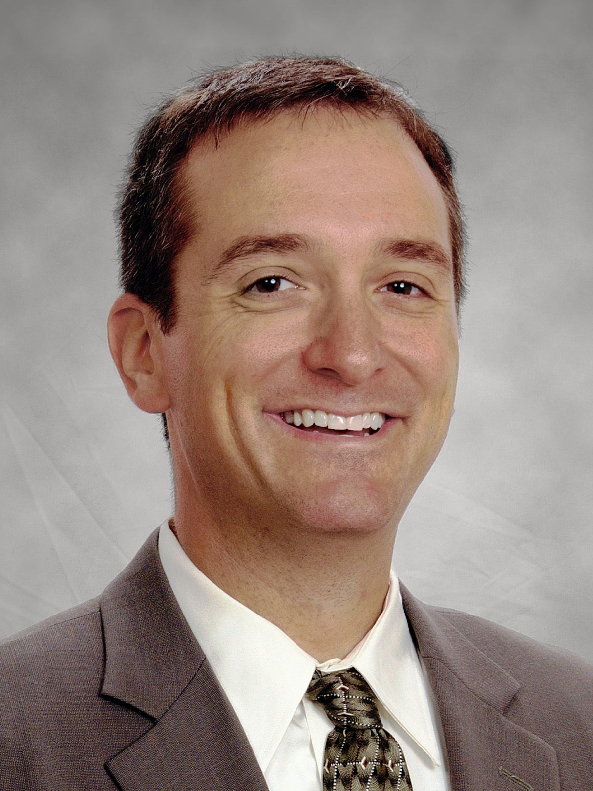 A man in a gray suit, white shirt, and patterned tie is smiling at the camera against a neutral gray background.