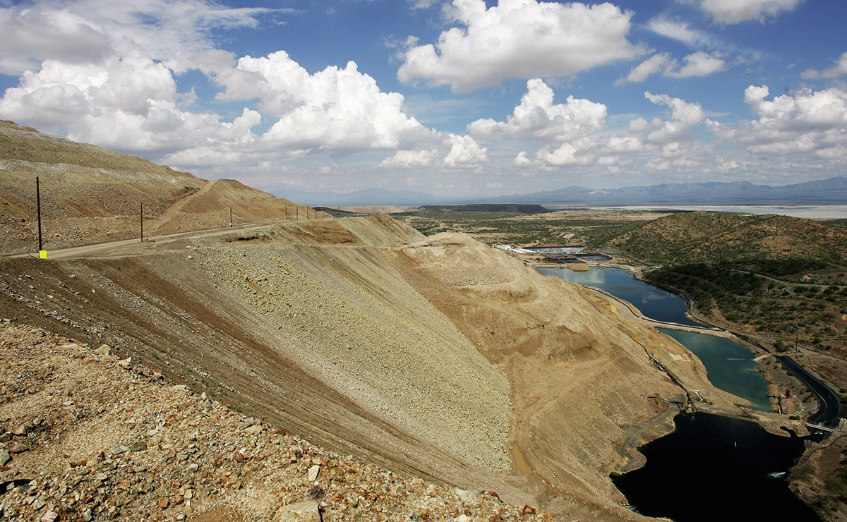 A large, terraced mining pit slopes down to several dark pools of water, surrounded by dry, rocky hills under a partly cloudy sky. Sparse vegetation appears on the hillsides in the background.