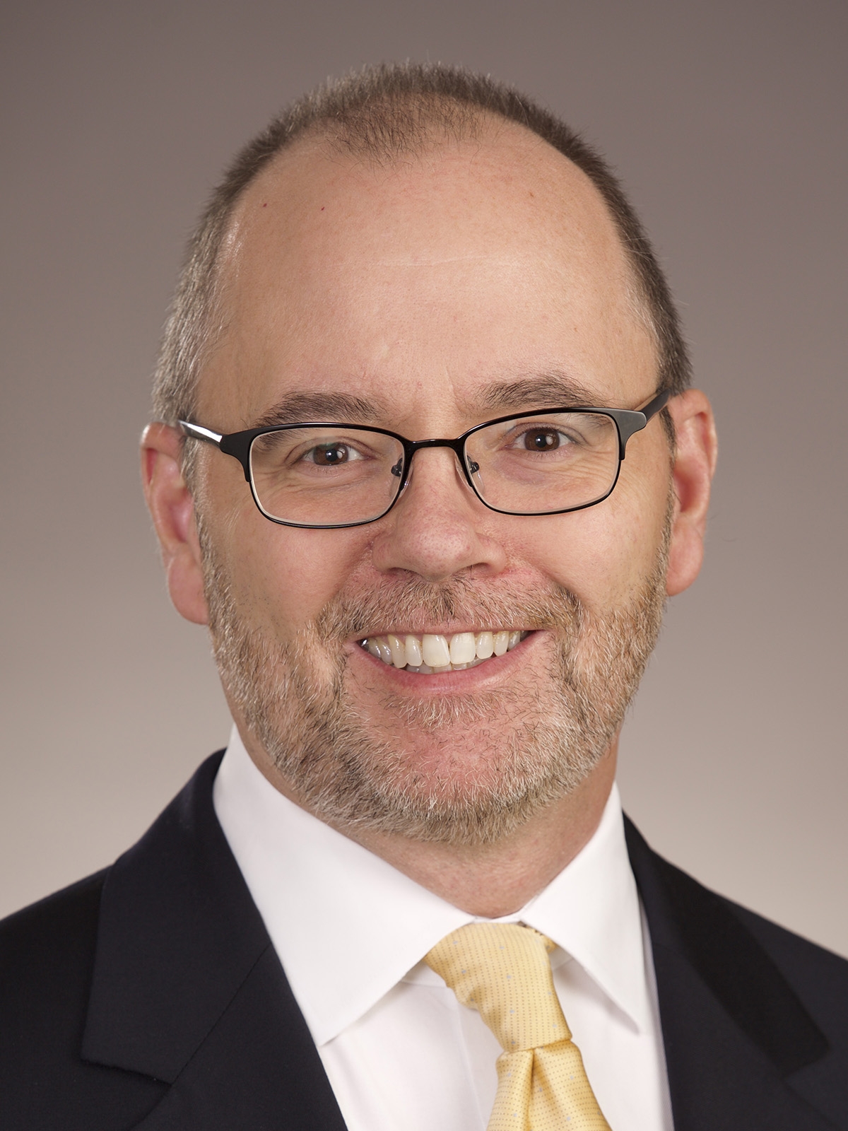 A middle-aged man with short, light brown hair, glasses, and a neatly trimmed beard, smiling and wearing a dark suit, white shirt, and yellow tie, posed against a plain, light background.