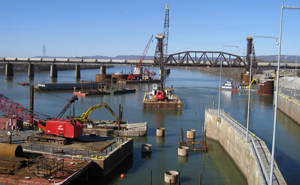 Construction equipment and cranes operate on barges in a river near a bridge, with concrete columns and a partially completed structure in the water. A lock or canal area is visible in the foreground.
