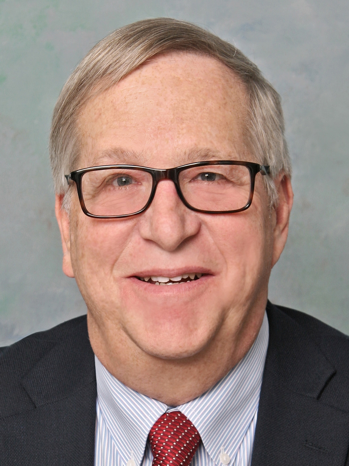A smiling older man with gray hair and glasses, wearing a dark suit, light blue shirt, and red patterned tie, posed in front of a light-colored background.