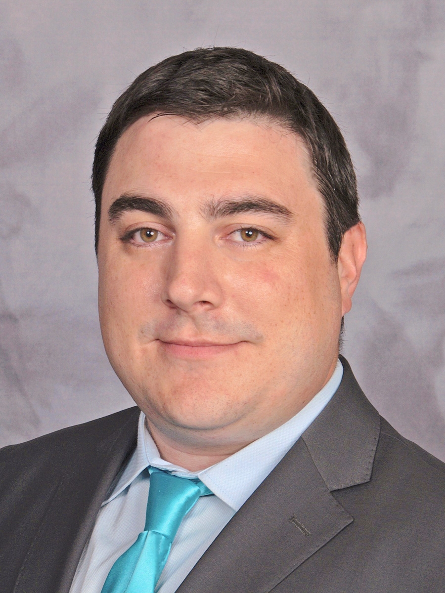 A man with short dark hair, light skin, and hazel eyes wearing a gray suit, light blue shirt, and turquoise tie, posed in front of a gray mottled backdrop.