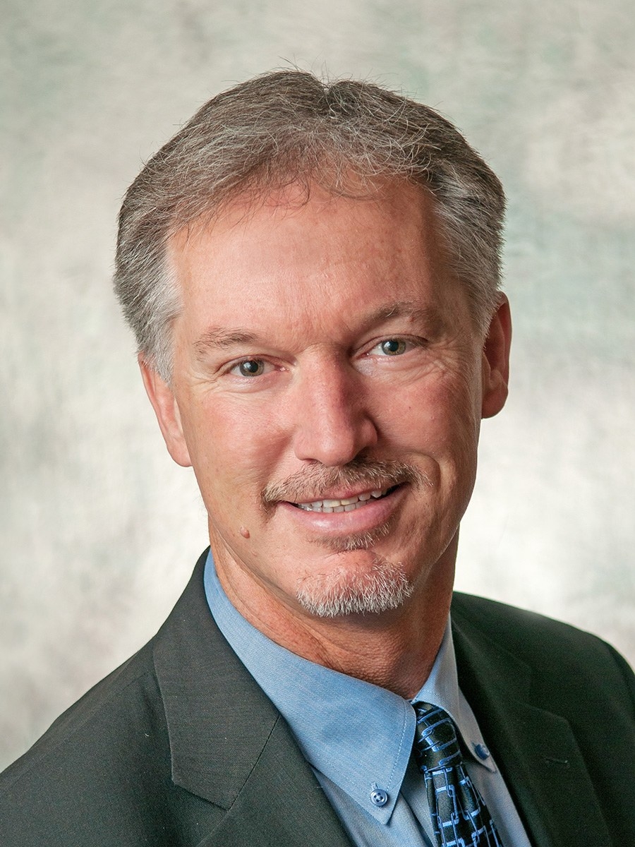 A middle-aged man with short gray hair and a trimmed goatee, wearing a dark suit jacket, blue dress shirt, and patterned tie, smiling at the camera against a light, textured background.