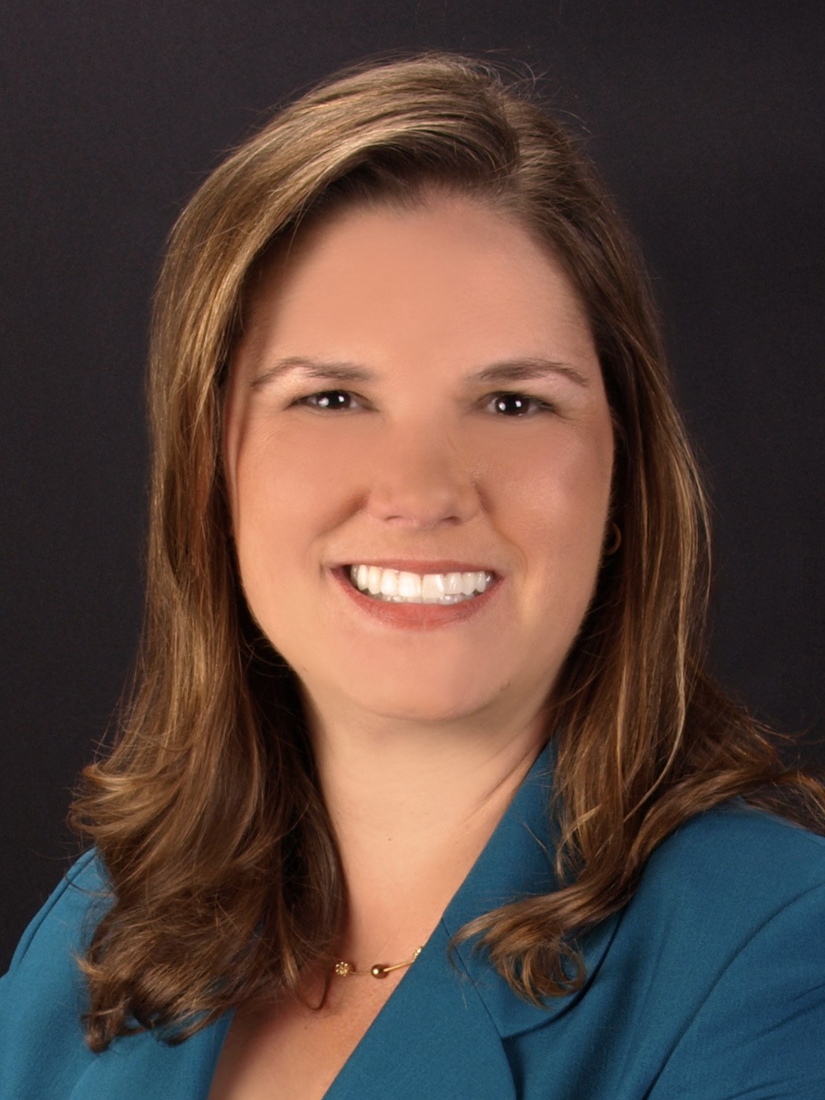 b262a56efdea6384d276521360f32e64 A woman with long, light brown hair is smiling at the camera. She is wearing a teal blazer and a gold necklace, posed against a plain dark background.