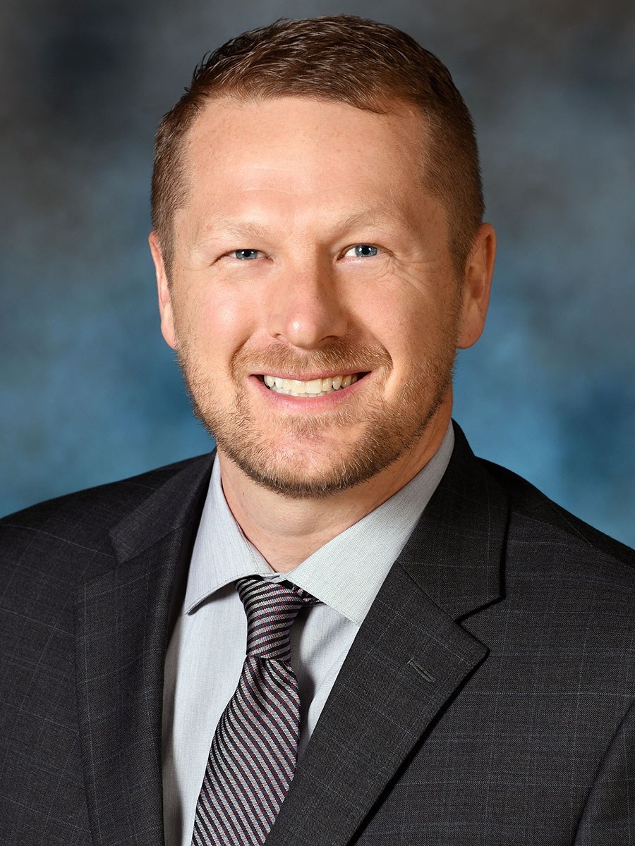 A man with short light brown hair and a trimmed beard, wearing a dark suit, light shirt, and striped tie, smiles at the camera in front of a blue and gray background.
