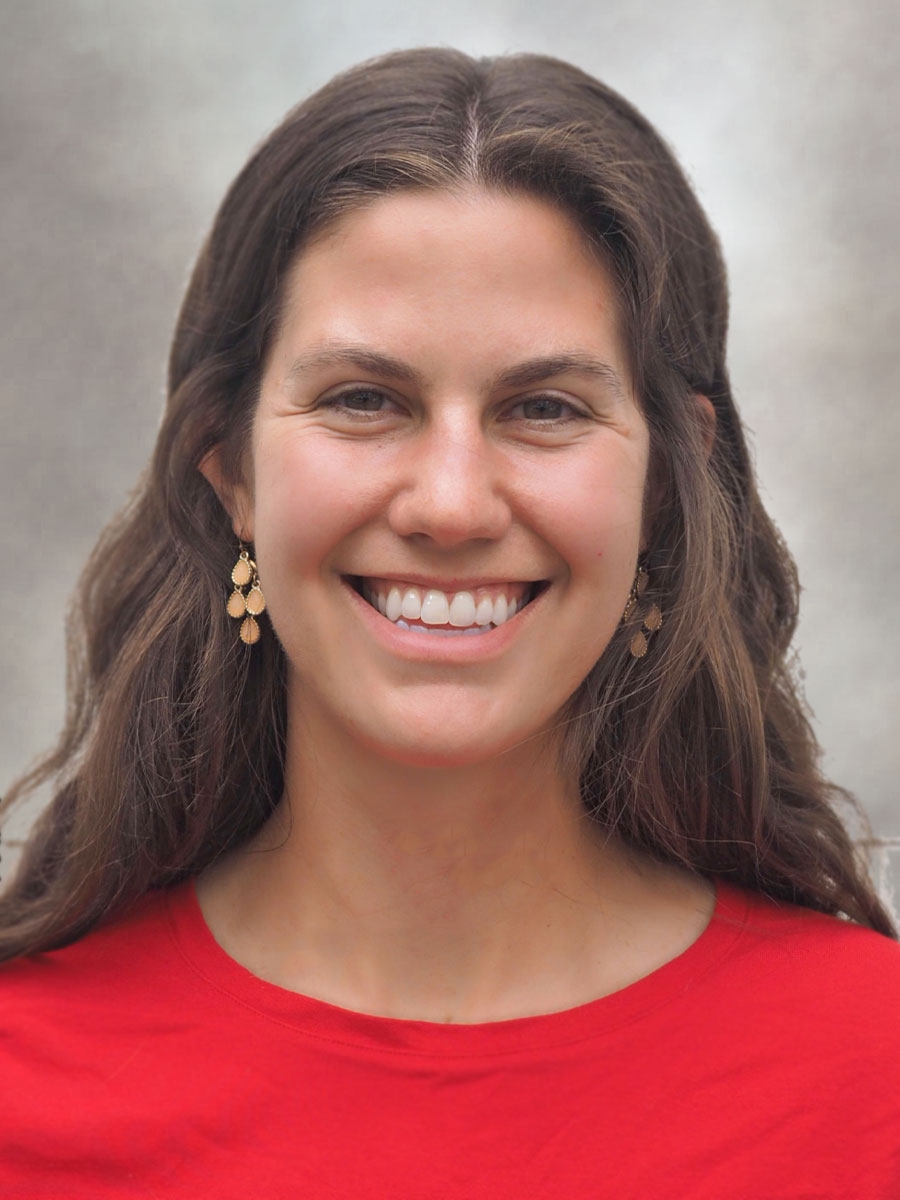 A woman with long brown hair, wearing gold drop earrings and a red top, smiles in front of a soft, neutral background.