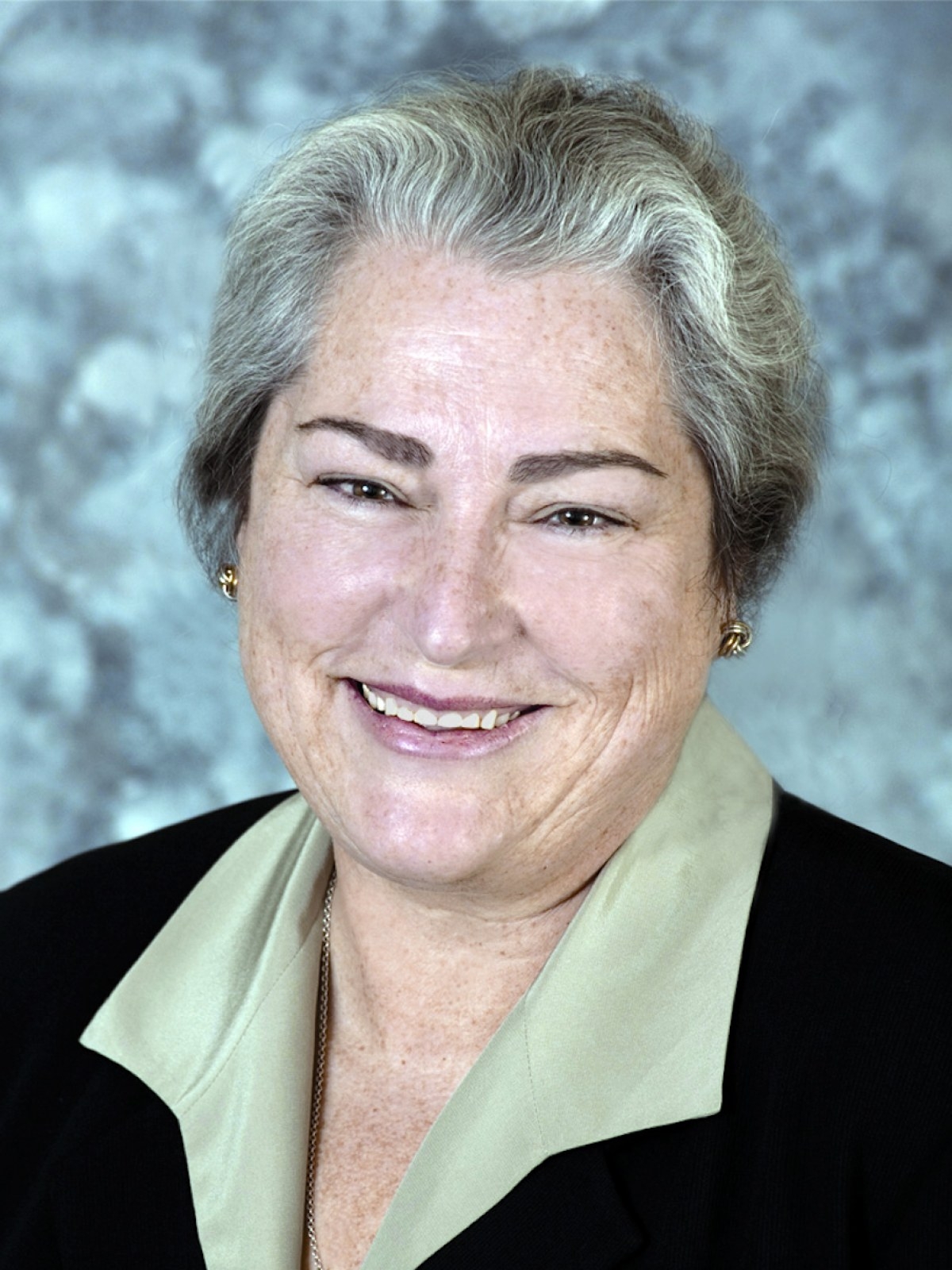 A middle-aged person with short gray hair smiles at the camera, wearing a black blazer over a light-colored blouse, with a blurred gray and white background.