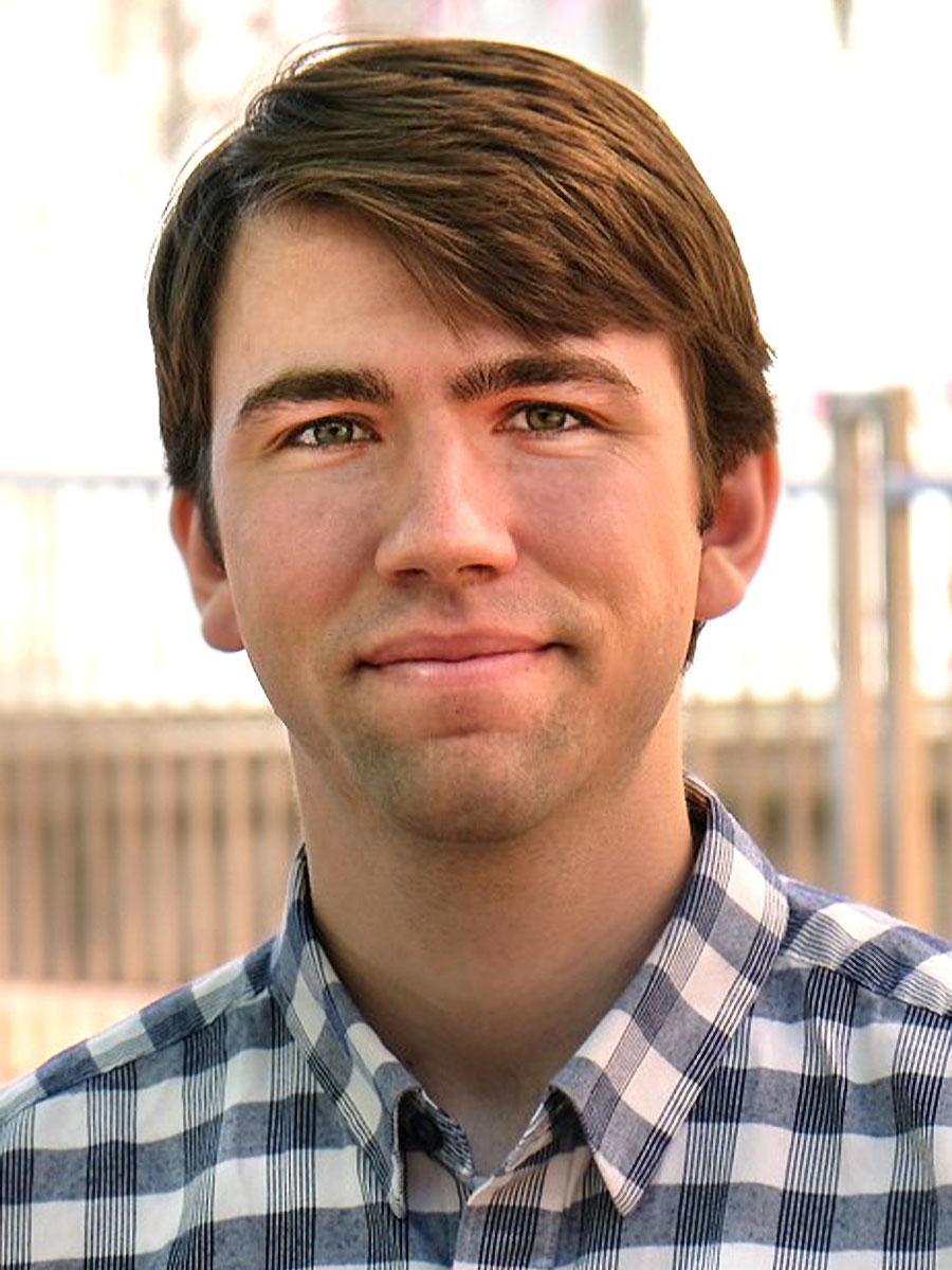 A young man with short brown hair and green eyes smiles gently at the camera. He is wearing a blue and white plaid shirt, and the background is softly blurred with neutral colors.