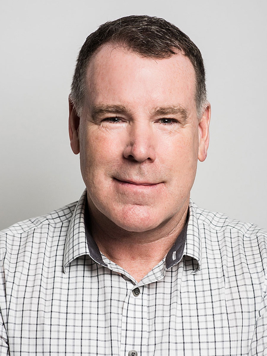 A middle-aged man with short brown hair, wearing a white plaid shirt, is smiling softly against a plain light gray background.