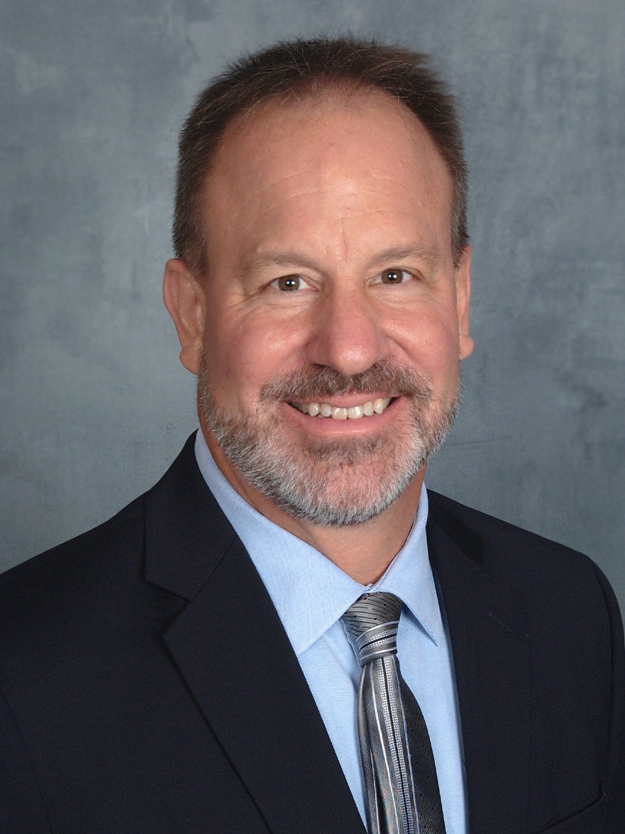 A man with short brown hair and a beard, wearing a dark suit, light blue shirt, and striped tie, smiles in front of a gray background.