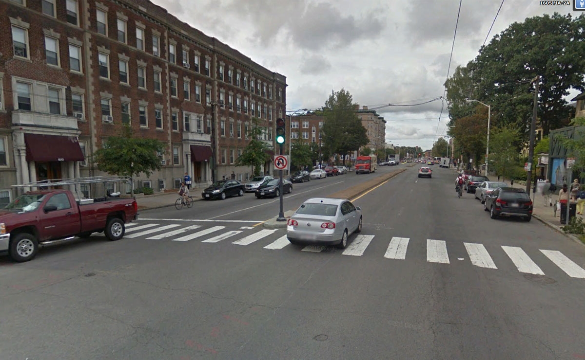 A city street intersection with cars stopped at a traffic light, a cyclist riding near the crosswalk, parked vehicles lining both sides, and multi-story brick buildings on the left under a cloudy sky.