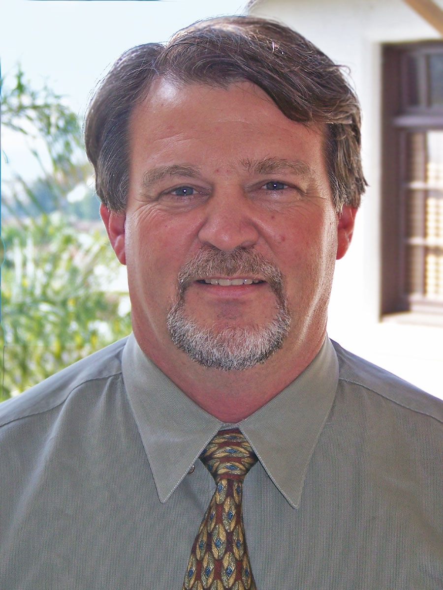 A middle-aged man with short brown hair, a mustache, and a salt-and-pepper goatee, wearing a light green dress shirt and patterned tie, stands indoors with greenery and a window in the background.