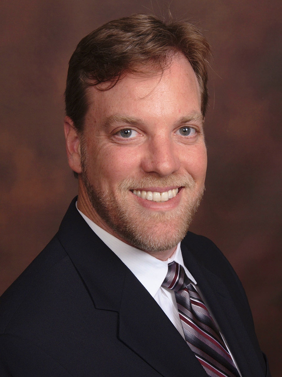 A man with light brown hair and a beard, wearing a dark suit, white shirt, and striped tie, smiling against a brown studio background.