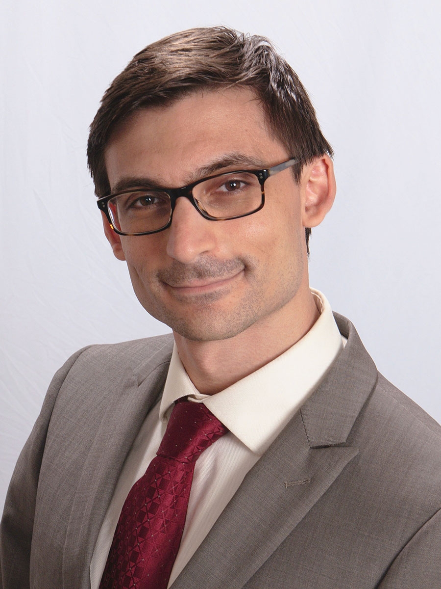 A person wearing glasses, a light-colored shirt, a grey suit, and a red patterned tie, smiling slightly while posing against a plain white background.