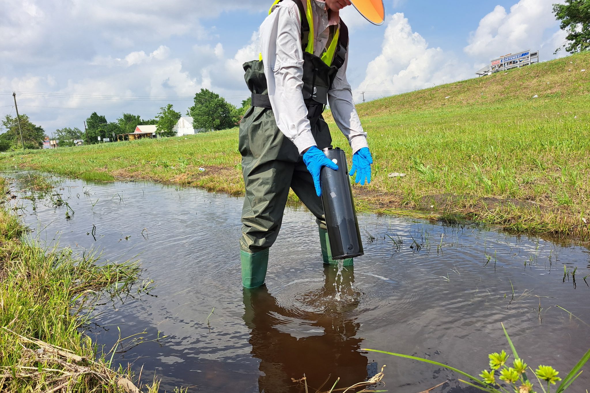 A person wearing waders, gloves, and a hat collects a water sample from a shallow stream or drainage ditch in a grassy area under a partly cloudy sky.