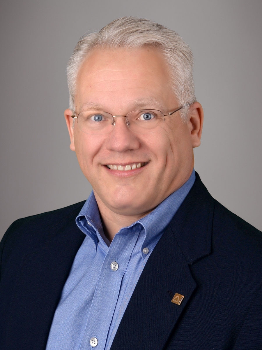 A smiling middle-aged man with short gray hair, glasses, and light skin, wearing a blue dress shirt and dark blazer, posing against a plain gray background.