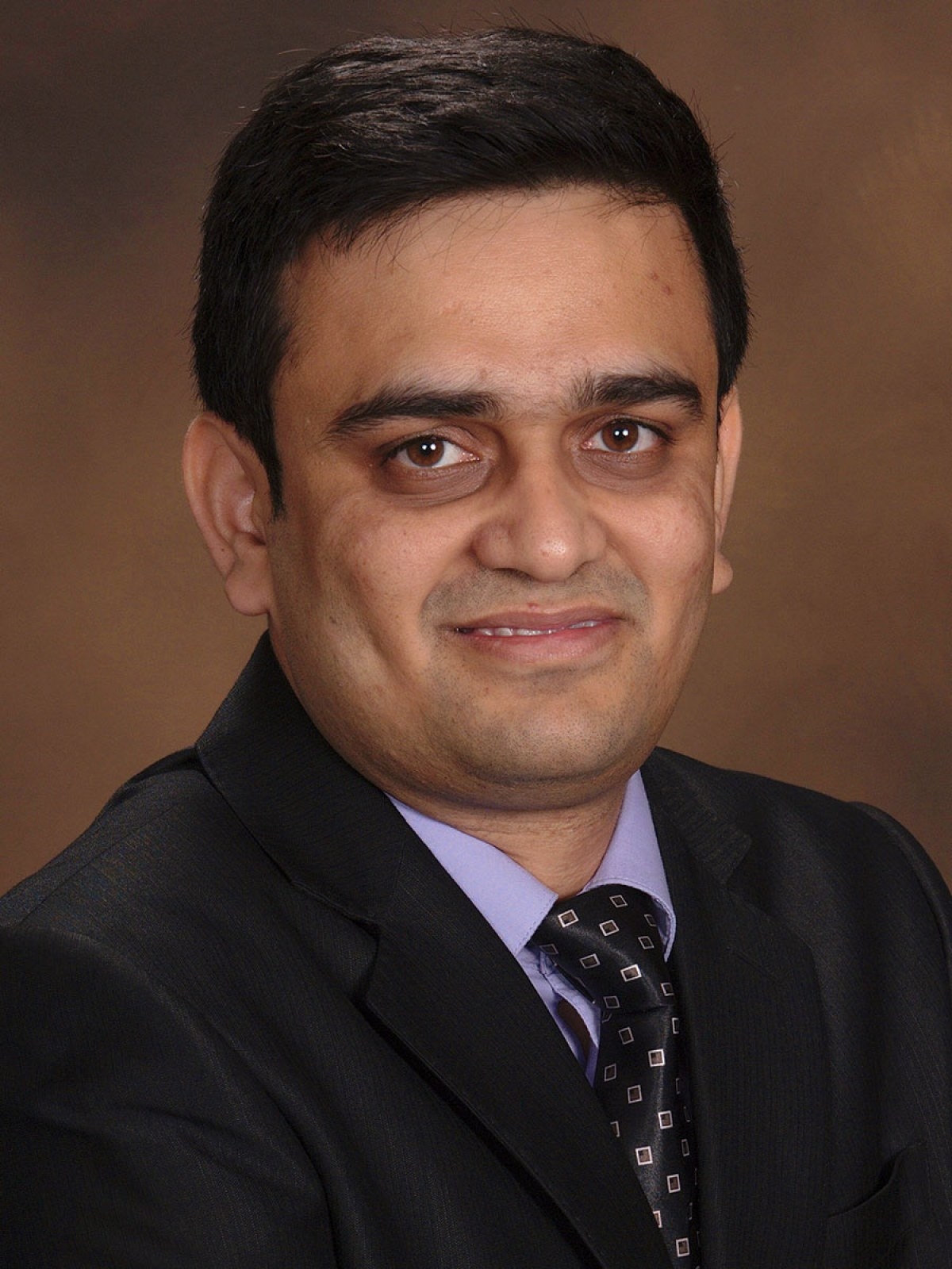 7a16e205791bc6643a6f360cda33ddba A man with short dark hair wearing a black suit, light purple shirt, and patterned tie smiles at the camera against a plain brown background.