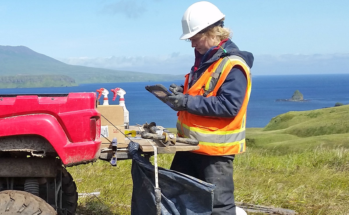 A person wearing a white hard hat, orange safety vest, and gloves uses a tablet near a red vehicle on grassy terrain, with the ocean and hills visible in the background.
