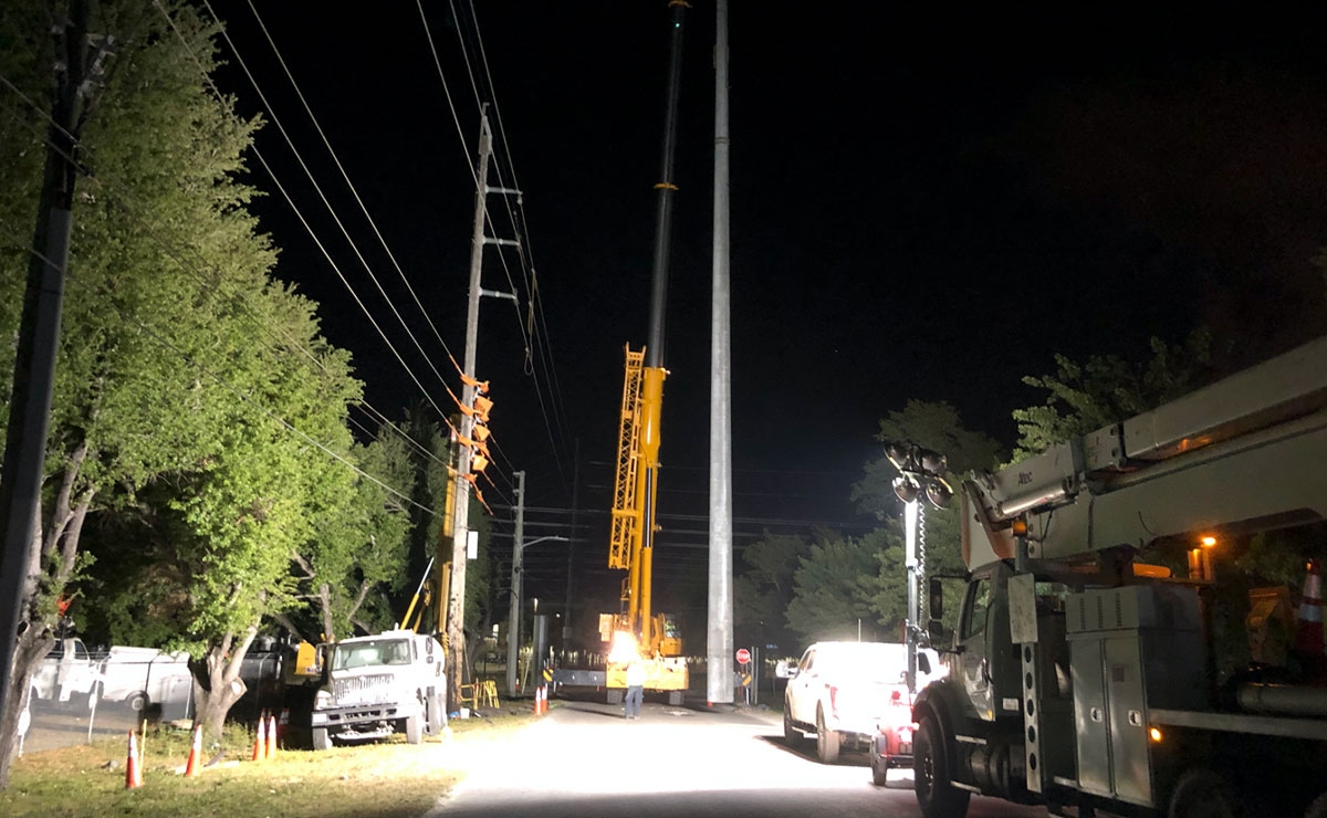A nighttime construction scene shows large utility trucks, workers in safety vests, and a tall crane illuminated by bright lights on a road lined with power lines and trees.