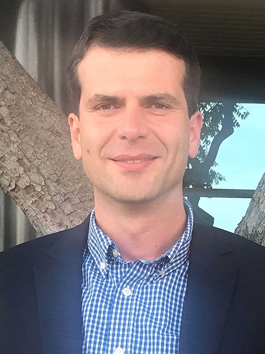 A man in a dark blazer and blue checkered shirt is smiling at the camera. He is standing outdoors in front of a tree and a building with reflective windows.
