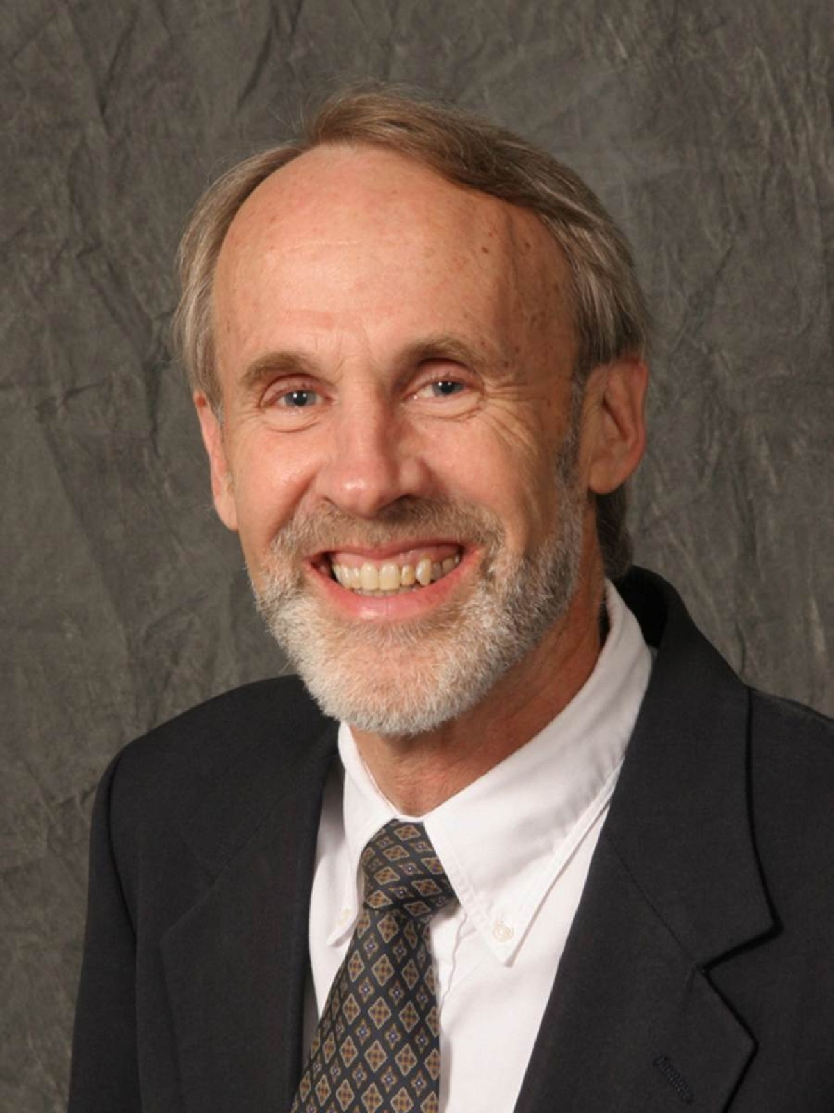 A smiling older man with a beard and short light hair wears a dark suit, white shirt, and patterned tie, posing in front of a textured gray background.