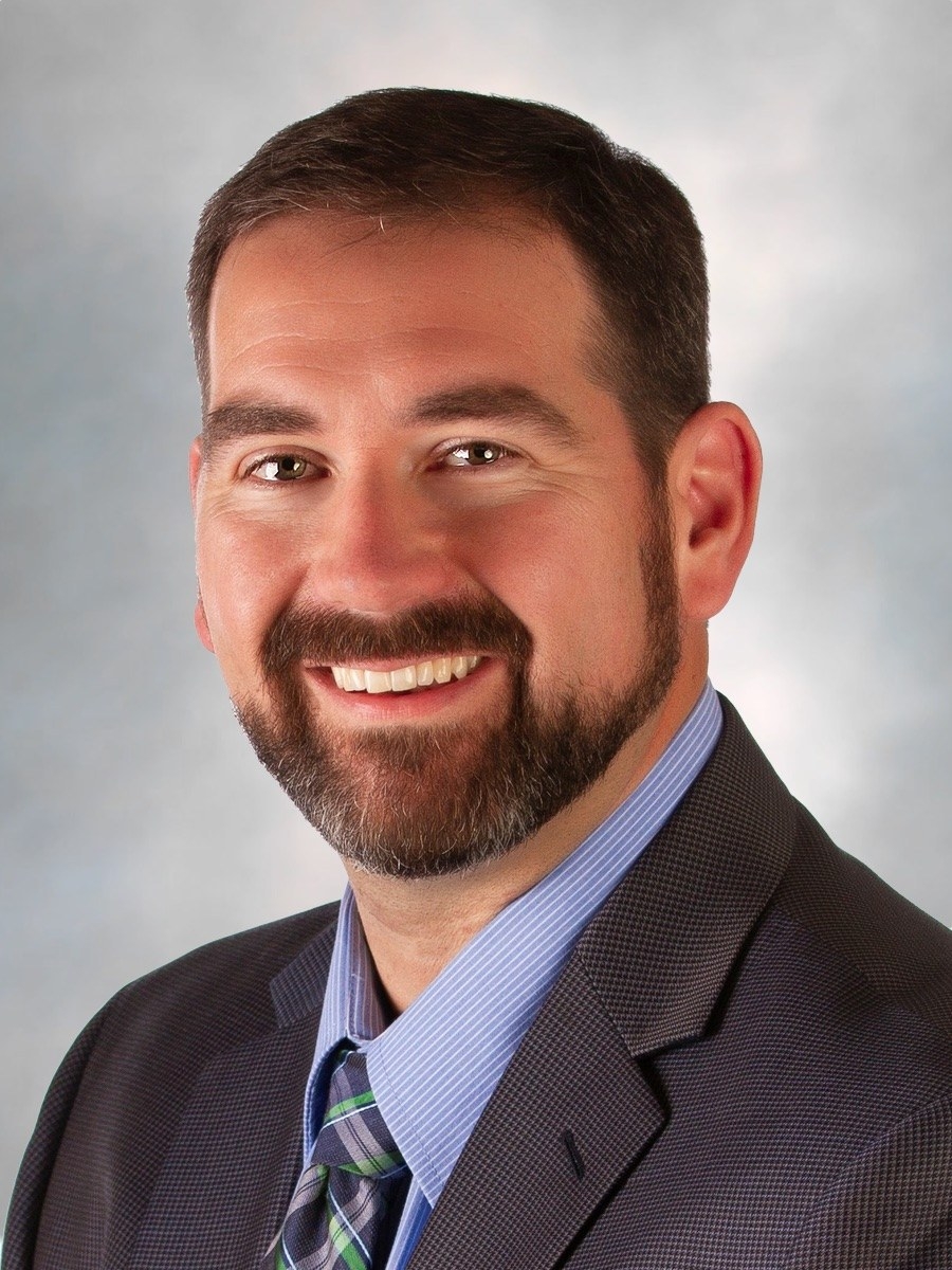 A man with short brown hair and a trimmed beard smiles, wearing a gray suit jacket, blue dress shirt, and striped tie, against a softly blurred light background.
