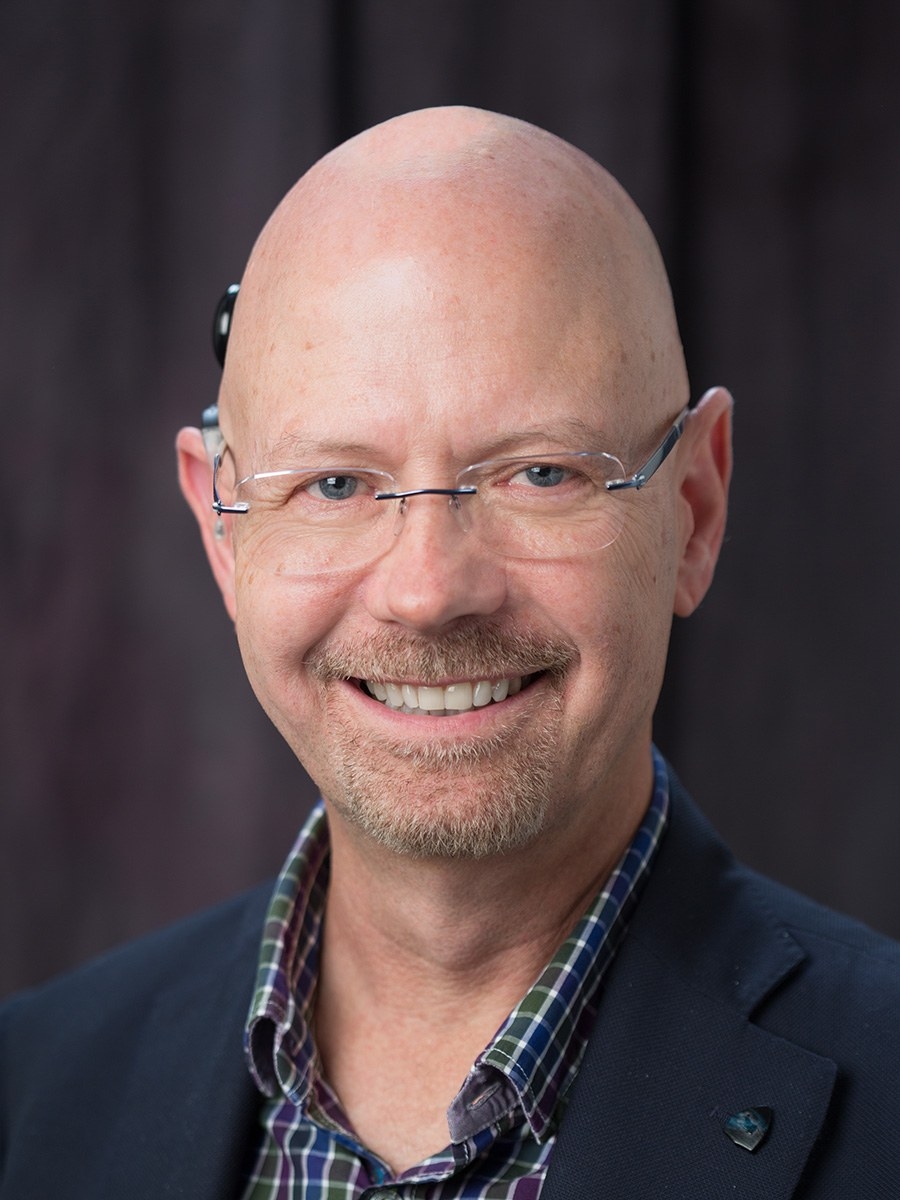 A smiling bald man with glasses and a goatee wears a plaid shirt and dark blazer, standing in front of a dark, blurred background.