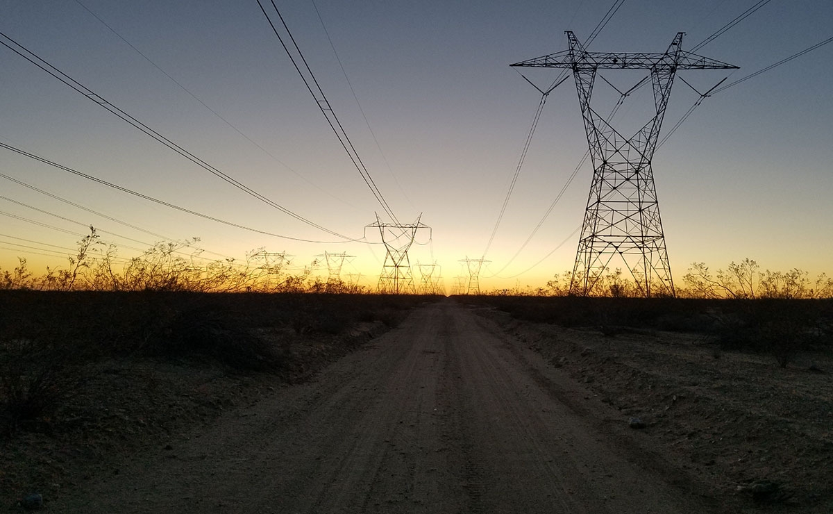 Dirt road running between tall power lines and towers at sunset, with silhouettes of sparse desert bushes on both sides and a clear sky in the background.