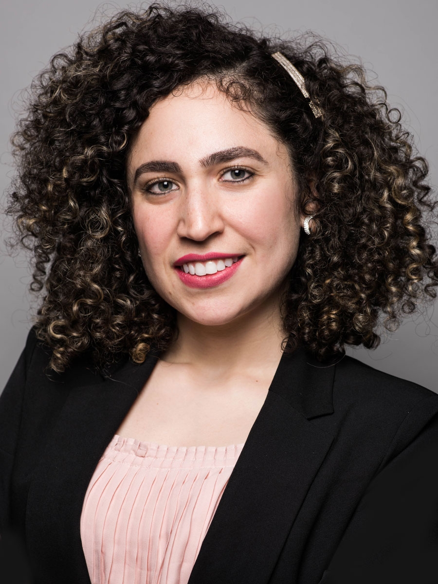 A woman with curly dark hair wearing a light pink blouse and black blazer smiles at the camera. She has a pearl earring and a hair clip, with a neutral gray background.