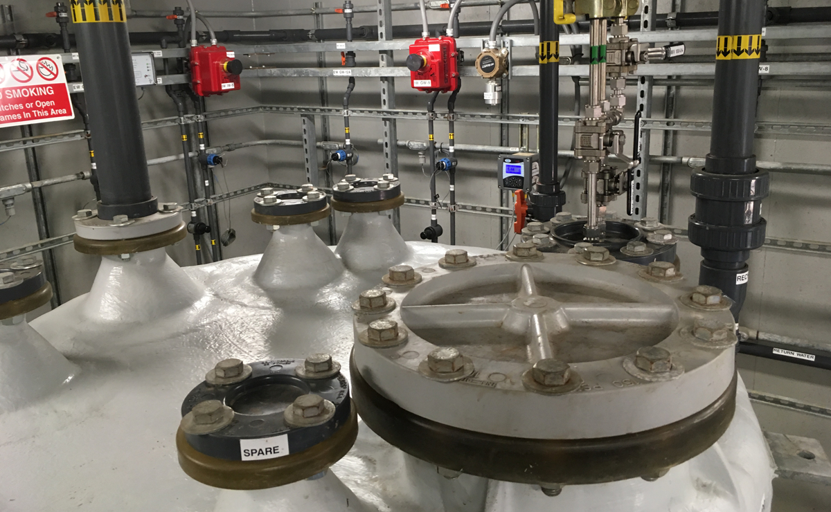 Large industrial pipes and valves with bolted flanges, labeled “SPARE,” in a control room featuring red control boxes, wiring, and safety signs on metal walls and conduit panels.